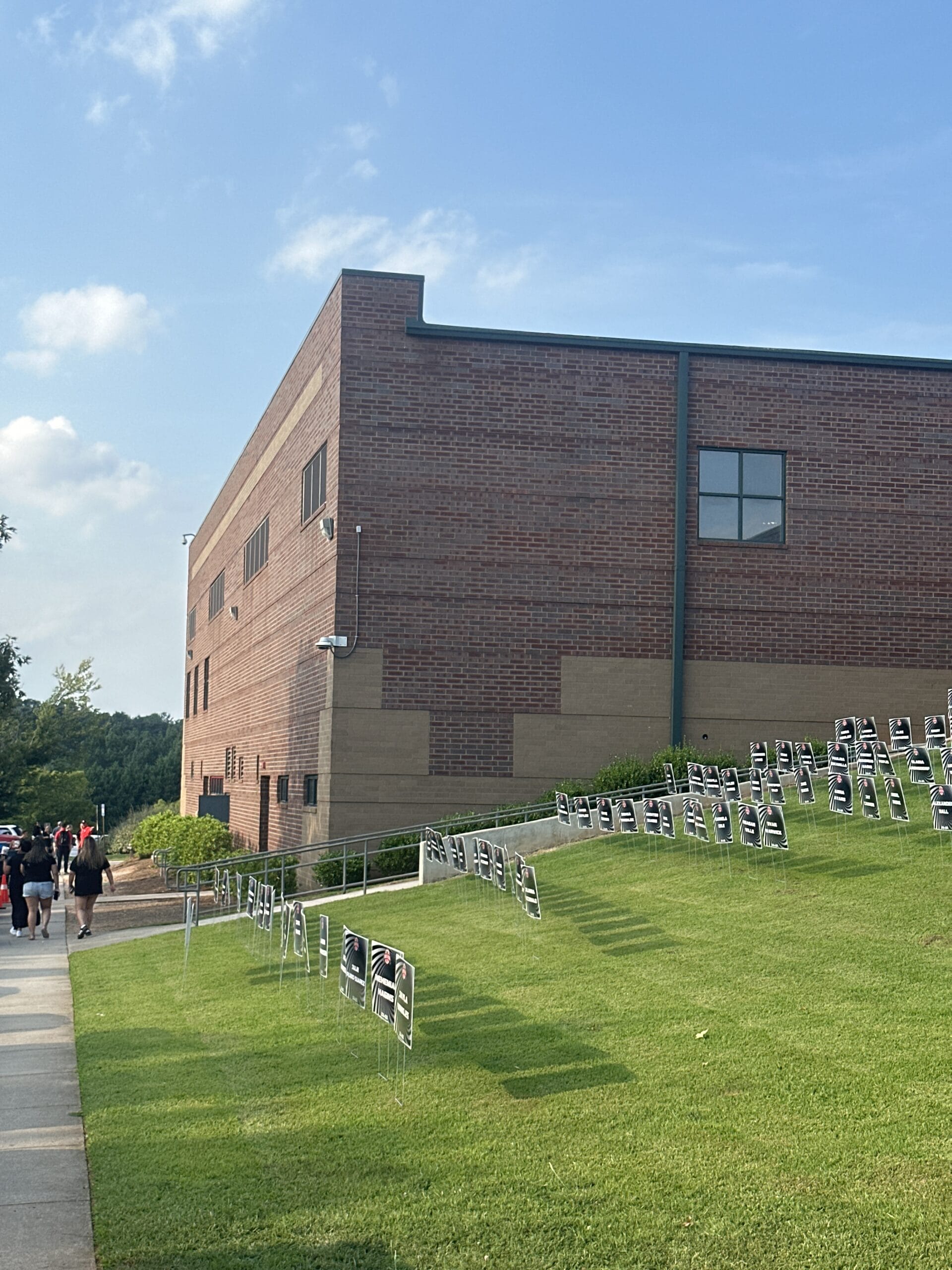 Side angle photo of a school with flyers on the lawn and people walking down the sidewalk to get to the event hidden from plain view.